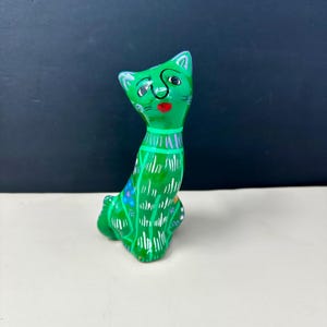May include: A green cat figurine with a whimsical design. The cat has a bright green body, a red mouth, and a collar with silver accents. The cat's body is decorated with white lines and colorful floral accents.