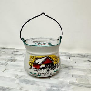 May include: A white ceramic savings bank with a black handle. The bank features a red barn scene with the words "Season's Greetings" and "Savings". The rim is green, and the handle is black.