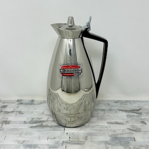 May include: A stainless steel insulated carafe with a black handle and lid. The carafe has a shiny, reflective surface and a label that reads "Qualität". The carafe is designed to keep beverages hot or cold.
