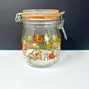 May include: A clear glass jar with an orange lid and metal clasp. The jar is decorated with a colorful pattern of vegetables, including onions, tomatoes, and artichokes. The jar is designed for food storage.