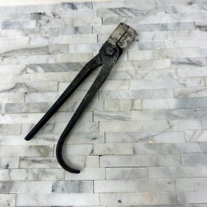 May include: A pair of heavy-duty, dark gray metal blacksmith tongs featuring a long, curved handle and a sturdy, rectangular jaw mechanism connected by a central pivot bolt. The tool exhibits a weathered, industrial finish with visible signs of use.