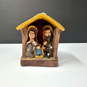 May include: A ceramic nativity scene featuring Mary, Joseph, and baby Jesus inside a rustic stable. The figures have halos and are painted with muted colors. The stable has a yellow roof and brown wooden walls.