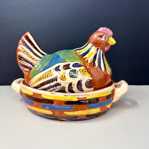 May include: A brightly painted ceramic chicken-shaped container. The chicken is brown with a pink comb and yellow beak, sitting in a matching bowl. The bowl and chicken are decorated with colourful stripes and patterns. The container has two small handles.
