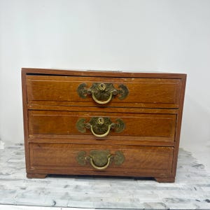 May include: A small, three-drawer wooden chest with brass hardware. The drawers are a warm brown color, and each has a decorative pull. The chest is likely used for storing small items, such as jewelry or trinkets.