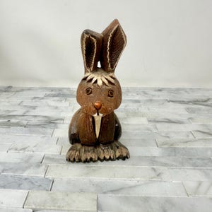 May include: A carved wooden rabbit figurine with a brown body, ears, and eyes. The rabbit has a white collar and a textured base. The figurine is a decorative item, possibly for Easter or spring.