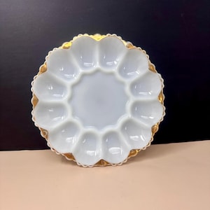 May include: A white, scalloped egg plate with a gold rim. The plate has 12 egg-shaped indentations around a central circular area. The gold rim adds a touch of elegance to the vintage serving dish.