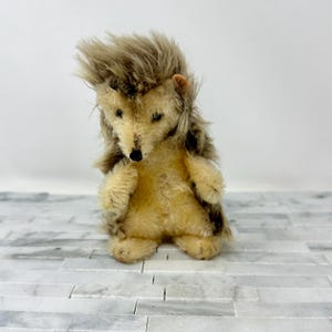 May include: A vintage stuffed hedgehog toy with a tan body, brown and tan fur, and a black nose. The hedgehog has small black eyes and is standing on a white and gray surface.