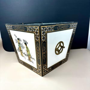 May include: A square, tapered wooden planter or box featuring a black finish with gold-painted floral borders. One side displays a painted scene of two figures in traditional attire, while the other side features a circular brass handle.