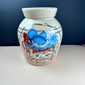 May include: A beige ceramic crock featuring a hand-painted blue bird sitting in a nest with red berries and a red ribbon. The text "His eye is on the sparrow. I know He watches me!" is written in black script around the upper portion of the vessel.