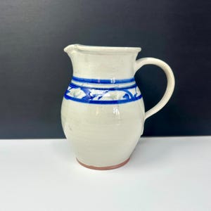 May include: A handmade ceramic pitcher with a white glaze and a blue floral design band. The pitcher has a curved handle and a spout for easy pouring. The base is a reddish-brown color.