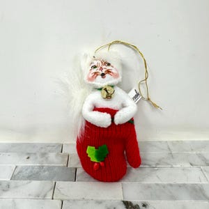 May include: A Christmas ornament shaped like a cat wearing a red mitten. The cat has a white face with rosy cheeks and a green collar with a small gold bell. The red mitten has a green holly leaf and berry design.
