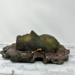 Puede incluir: Escultura de cer&aacute;mica de un rostro humano en tonos verdes, marrones y dorados. El rostro descansa sobre una base texturizada de forma org&aacute;nica. Un cord&oacute;n dorado est&aacute; unido a la escultura. La obra de arte se exhibe sobre una superficie blanca y gris.