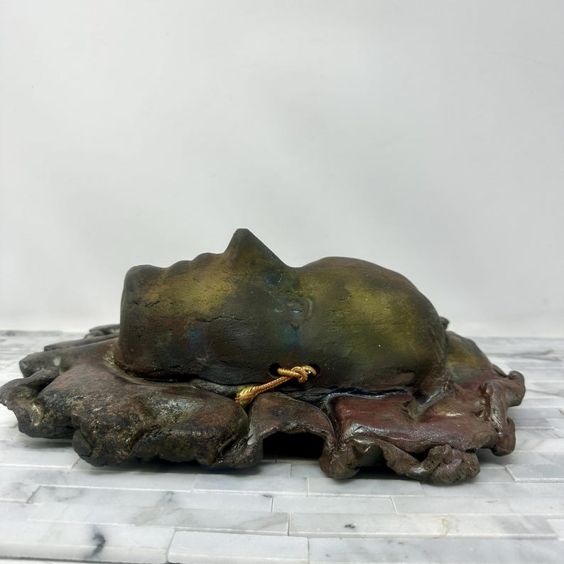 Puede incluir: Escultura de cer&aacute;mica de un rostro humano en tonos verdes, marrones y dorados. El rostro descansa sobre una base texturizada de forma org&aacute;nica. Un cord&oacute;n dorado est&aacute; unido a la escultura. La obra de arte se exhibe sobre una superficie blanca y gris.