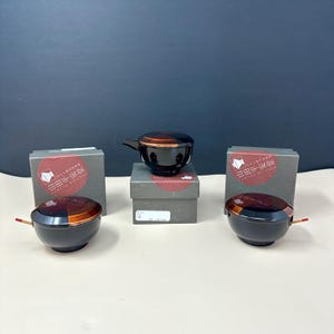 May include: Black and brown lacquered bowls with lids and a handle, displayed with their original packaging. The bowls have a glossy finish with a copper-colored accent. The packaging features Japanese text and a red circular design.
