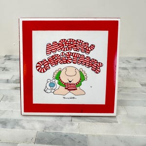 May include: A square, red-framed Christmas decoration with a white center. The center features a cartoon illustration of a character wearing a green shirt and red scarf, with a small blue dog. The words "MERRY CHRISTMAS" are written in red and white candy cane style.