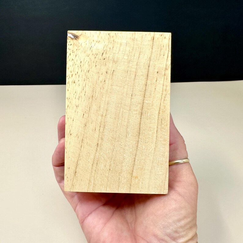 Op de afbeelding: Een rechthoekig blok lichtgekleurd hout met zichtbare nerven en kleine donkere vlekken. Het houten blok wordt in een hand gehouden, met een gouden ring zichtbaar op een vinger. De achtergrond is een mix van zwart en gebroken wit.