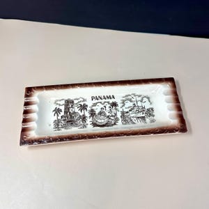 May include: Rectangular ceramic ashtray with a brown border and a white interior. The ashtray features a black and white illustration of Panama, including the word "PANAMA" in the center. The design includes images of buildings, people, and ships.