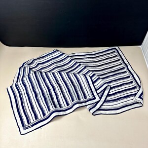 May include: A rectangular scarf featuring a white background with navy blue vertical and horizontal stripes. The fabric has a smooth texture and a finished border. A small white rectangular tag is attached to one corner with visible text.