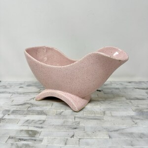 May include: A pink ceramic planter with a speckled finish. The planter has a curved, boat-like shape and sits on a curved base. The interior is smooth, and the exterior has a textured appearance.