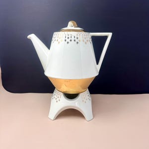 May include: A vintage white and gold teapot with a matching warmer. The teapot features a geometric design with gold accents and a gold base. The warmer is white with gold starburst patterns.