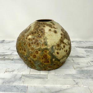 May include: A handcrafted, organic-shaped ceramic vase. The vase features a speckled pattern with shades of brown, green, and beige. The opening is a dark brown color. The vase is a decorative item for the home.