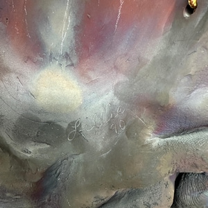Puede incluir: Primer plano de una escultura texturizada con tonos grises, morados y blancos. La firma del artista, "Lillie '90", est&aacute; grabada en la superficie. La superficie de la escultura tiene un aspecto rugoso y org&aacute;nico. La obra de arte tiene un aspecto multicolor.