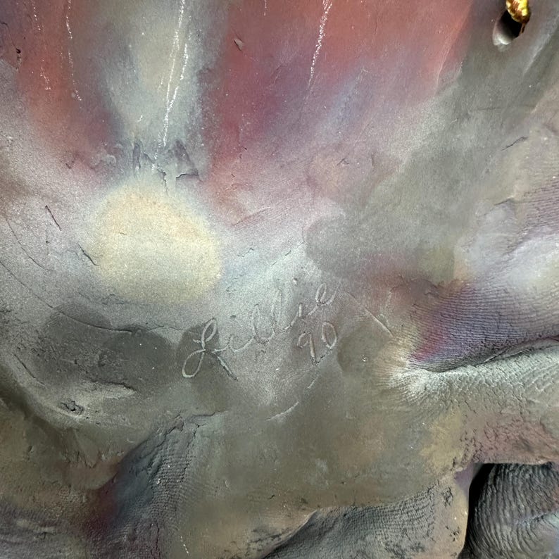Puede incluir: Primer plano de una escultura texturizada con tonos grises, morados y blancos. La firma del artista, "Lillie '90", est&aacute; grabada en la superficie. La superficie de la escultura tiene un aspecto rugoso y org&aacute;nico. La obra de arte tiene un aspecto multicolor.