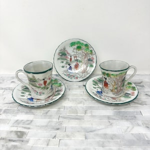 May include: A set of three vintage teacups and saucers. The cups and saucers feature a hand-painted Asian-inspired landscape design with figures, trees, and buildings. The rims are trimmed with green. The set includes two cups and saucers and one additional saucer.