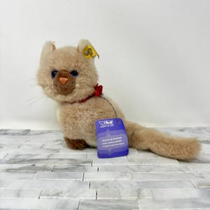 May include: A plush tan cat toy with blue eyes, a red collar, and a small red bow. The cat has a brown nose and paws. A blue tag with white text is attached to the toy. The cat is lying on a white and gray surface.
