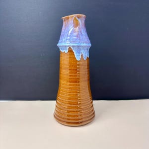 May include: A handmade ceramic jug with a brown, ribbed body and a flared top. The top is glazed in shades of blue and purple, creating a unique, decorative piece. The jug has a spout for easy pouring and is suitable for serving beverages.