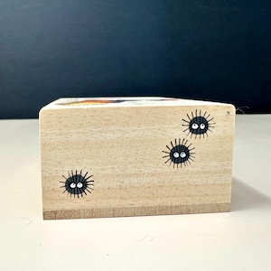 Op de afbeelding: Een kleine, rechthoekige houten blok met handgeschilderde zwarte roetgeesten. Elke geest heeft een rond zwart lichaam met witte ogen en een kleine, lachende mond. Het blok is gemaakt van lichtgekleurd hout.