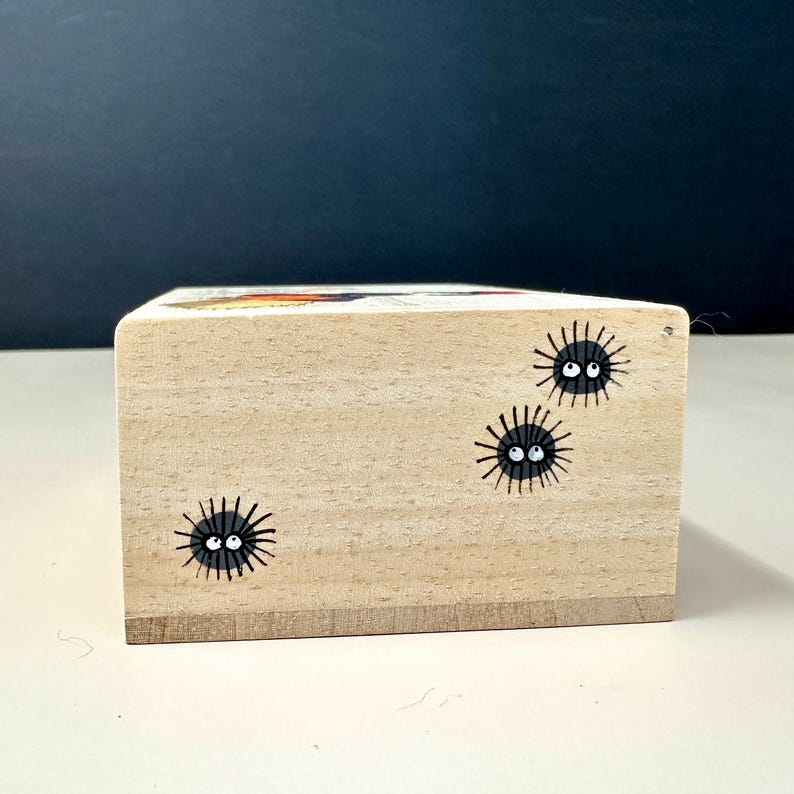Op de afbeelding: Een kleine, rechthoekige houten blok met handgeschilderde zwarte roetgeesten. Elke geest heeft een rond zwart lichaam met witte ogen en een kleine, lachende mond. Het blok is gemaakt van lichtgekleurd hout.