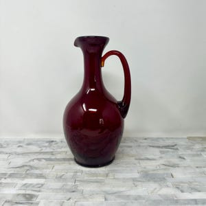 May include: A tall, burgundy-colored glass pitcher with a rounded body and a curved handle. The pitcher has a spout for pouring and is likely used for serving beverages. The glass has a glossy finish.
