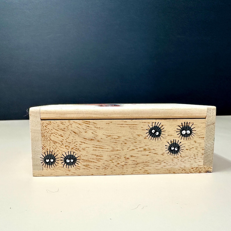 Op de afbeelding: Een kleine, rechthoekige houten doos met een lichte houtnerf afwerking. De doos is versierd met zwarte, stekelige, cartoonachtige wezens met witte ogen. De doos heeft een deksel en staat op een wit oppervlak.