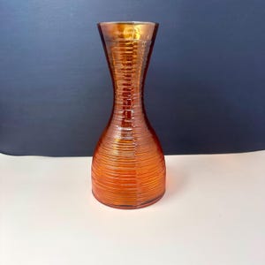 May include: An orange glass vase with a textured, ribbed design. The vase has a slender, hourglass shape, widening at the base and top. The glass has a warm, amber hue, catching the light and creating a shimmering effect.