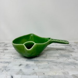 May include: A bright green ceramic serving dish with a handle. The dish has a unique shape with a central, smaller compartment and a larger, open area. The handle is rectangular with a textured design. The dish is on a white and gray surface.