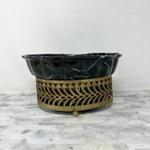 May include: A black and white marble-patterned bowl with a scalloped edge sits atop a gold-colored, ornate metal base. The base has a decorative, openwork design. The bowl and base are likely for decorative or functional use.