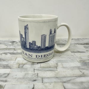 May include: White ceramic mug featuring a blue and white illustration of the San Diego skyline. The mug has a curved handle and the words "SAN DIEGO AMERICA'S FINEST CITY" are printed on the bottom.