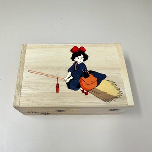 Op de afbeelding: Houten kist met een illustratie van een jonge heks op een bezem. De heks draagt een donkerblauwe jurk, een rode strik en een oranje tas. De kist heeft een lichte houtnerf afwerking en sterdecoraties.
