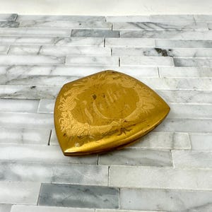 May include: A vintage, gold-toned compact with a floral and leaf design. The compact has a heart-shaped design with the word "thee" in an oval in the center. The compact is on a white and gray marble surface.