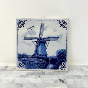May include: Square ceramic tile featuring a blue and white illustration of a Dutch windmill. The windmill is set against a cloudy sky and surrounded by foliage. The word "Holland" is written in script. Decorative scrollwork borders the image.