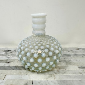May include: A clear glass vase with a round body and a short, cylindrical neck. The body is covered in small, white, raised dots, giving it a textured appearance. The neck is a solid white color. The vase is sitting on a light-colored surface.