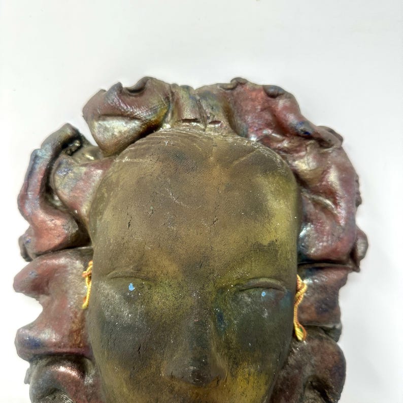Puede incluir: Una escultura de rostro de cer&aacute;mica con una apariencia desgastada y envejecida. La cara es principalmente marr&oacute;n con toques de verde y azul, y el cabello es una mezcla de rojo y morado. Pendientes dorados cuelgan de las orejas.