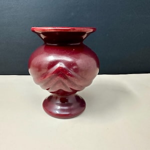 May include: A burgundy-colored ceramic vase with a rounded body and a flared rim. The vase has a textured, carved design on the lower half and sits on a circular base. The background is a gradient of white and black.