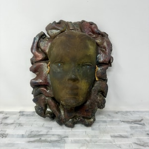 Puede incluir: Escultura mural en tonos bronce de un rostro humano estilizado con pendientes dorados. El rostro est&aacute; enmarcado por un dise&ntilde;o abstracto texturizado y multicolor. La escultura tiene un aspecto envejecido, con toques de verde y rojo.