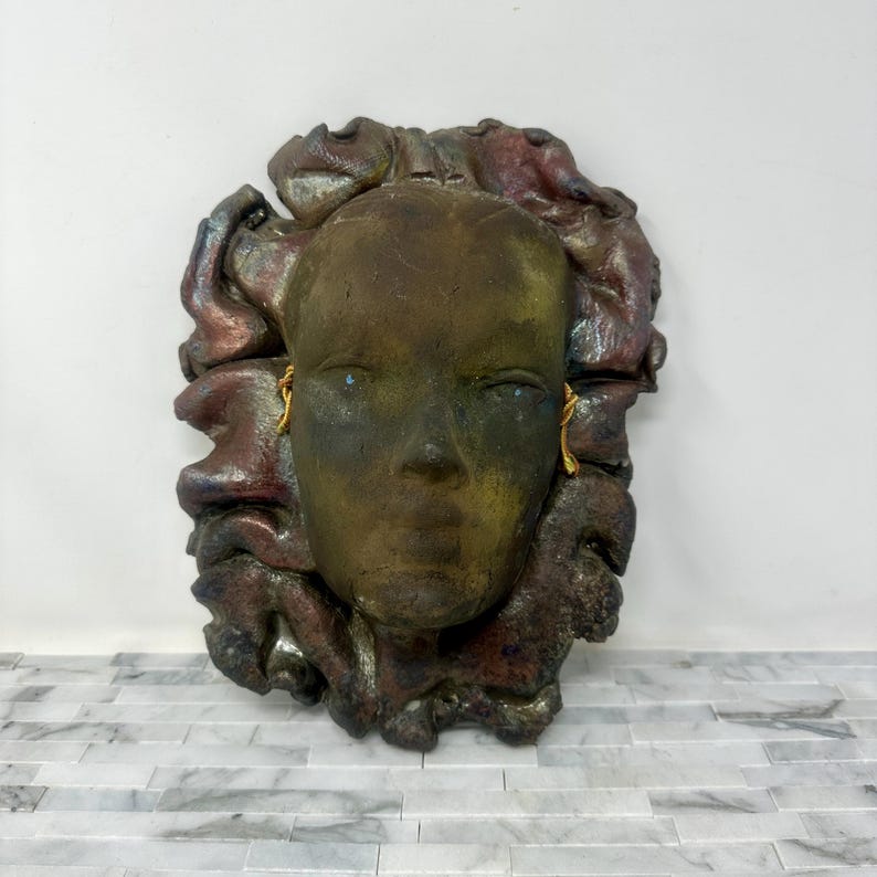 Puede incluir: Escultura mural en tonos bronce de un rostro humano estilizado con pendientes dorados. El rostro est&aacute; enmarcado por un dise&ntilde;o abstracto texturizado y multicolor. La escultura tiene un aspecto envejecido, con toques de verde y rojo.