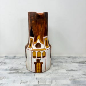 May include: A tall, decorative ceramic pitcher with a church-like design. The pitcher features a white base with yellow and brown accents, and a brown top. The design resembles a building with arched windows and a central door.