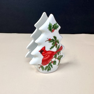 May include: White ceramic Christmas tree-shaped napkin holder. The holder features a red cardinal bird perched on a holly branch with red berries. The design is repeated on both sides of the holder.