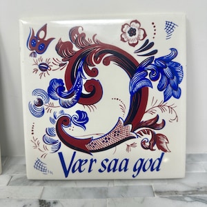 May include: A white ceramic tile with a blue and red floral design. The tile features a stylized letter "D" and the text "Vaer saa god".