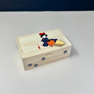 Op de afbeelding: Een lichtgekleurde houten kist met een cartoonillustratie van een jonge heks op een bezem. De kist heeft een scharnierend deksel en is versierd met kleine, zwarte, oogachtige wezens. De heks draagt een blauwe jurk en een oranje tas.